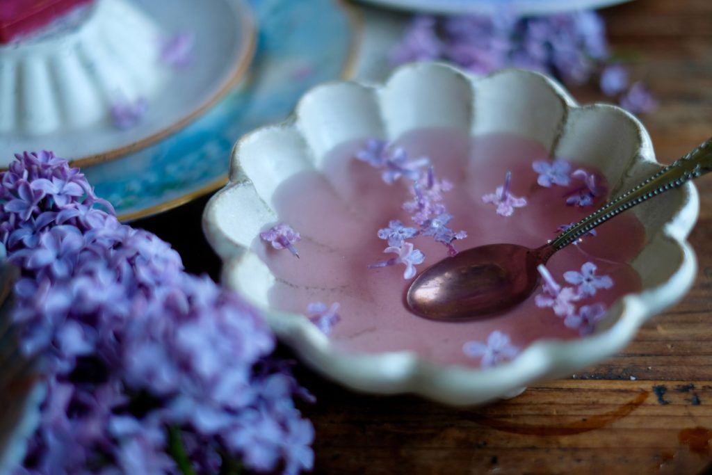 lilac syrup in a bowl with a spoon surrounded by lilac flowers